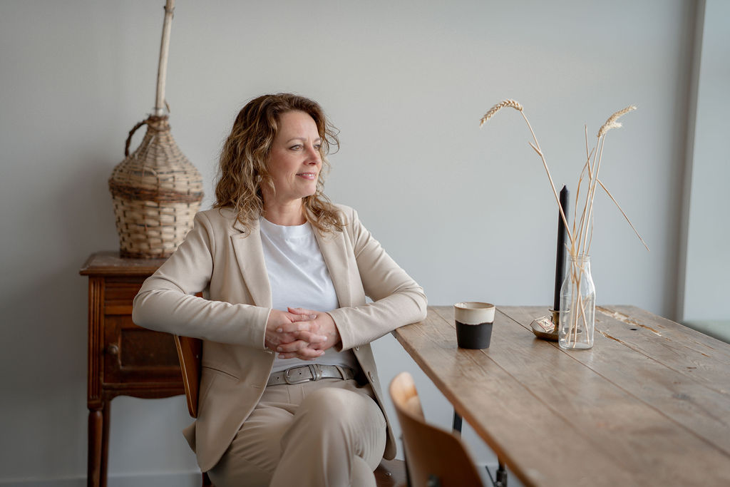 Jannie de Jong - Virtueel assistent. Zittend aan de tafel, naar buiten kijkend. Met de handen in elkaar.