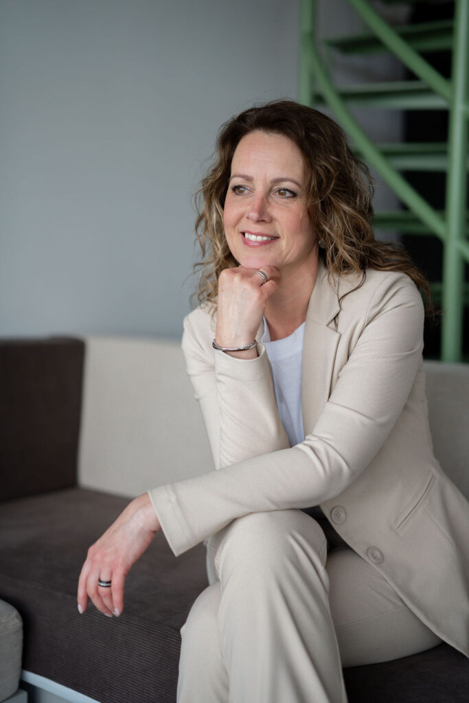 Jannie de Jong met beige pak aan met hand onder kin en glimlachend