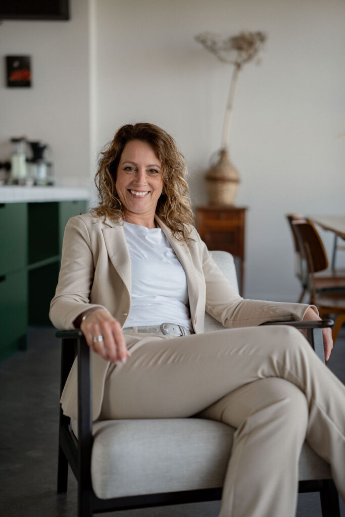Virtueel assistent Jannie zittend en glimlachend in stoel met beige pak aan.